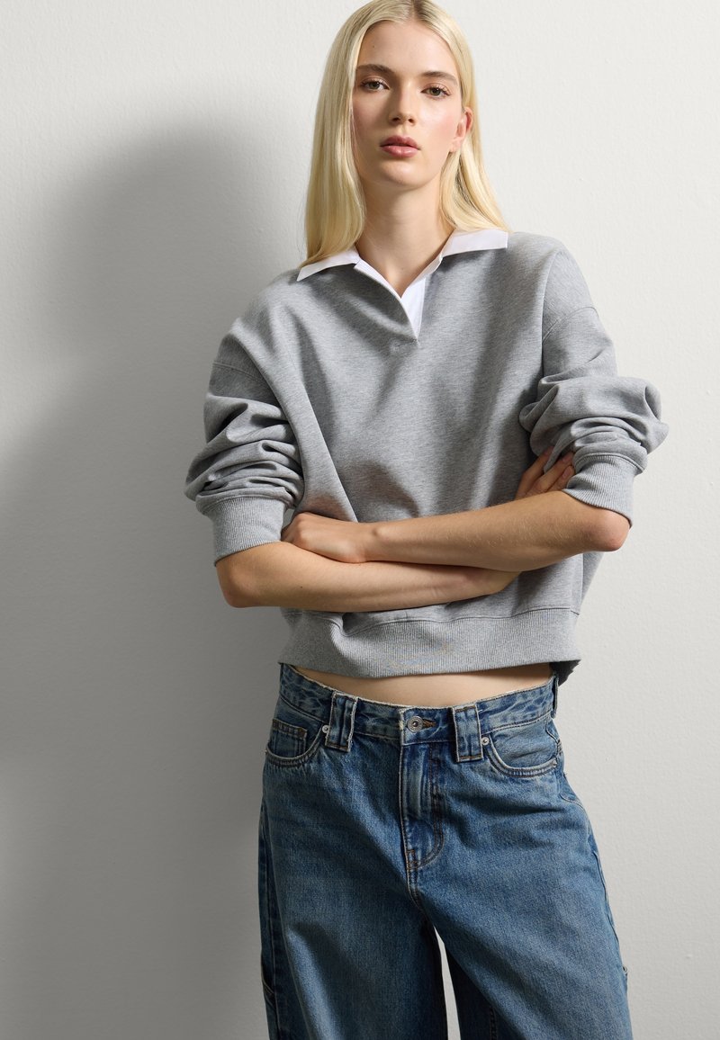 Grijze cropped sweatshirt met een witte kraag, gecombineerd met blauwe oversized jeans. De stof lijkt zacht, met een losse pasvorm en opgerolde mouwen.