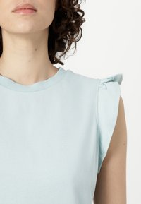 Hele sinine ilma varrukateta topp, mis on valmistatud puuvillast, ringatrahvi ja kogutud kangakatete detailidega õlgadel. Sile tekstuur.