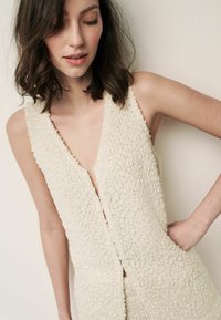 Combinaison en tricot crème texturée avec un décolleté en V profond, sans manches et poches latérales. Le matériau semble doux et légèrement pelucheux.