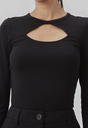 Femme portant un haut noir à manches longues avec une découpe en forme de goutte à l'encolure et un pantalon noir taille haute avec fermeture boutonnée.