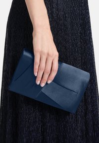 Pochette en satin bleu marine avec un design géométrique plié, tenue dans une main, sur un fond textur é sombre.