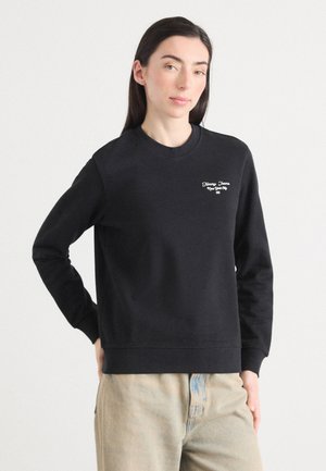 Ung kvinna med långt mörkt hår klädd i en svart sweatshirt och ljusa byxor, poserar mot en enfärgad vit bakgrund.