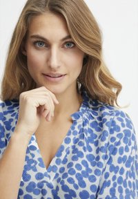 Blusa blu e bianca con un motivo a fiori discreto, caratterizzata da un scollo a v e colletto ruffled, indossata da una persona con capelli mossi.