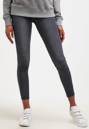 Personne portant un jean slim gris foncé et des baskets blanches, debout, jambes écartées, sur un fond clair uni.