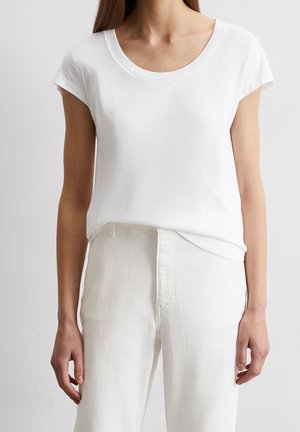 Femme portant un haut blanc uni à encolure dégagée et manches courtes, rentré dans un pantalon blanc taille haute, debout devant un fond clair uni.