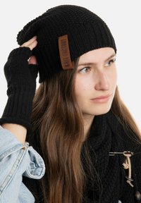 Zwarte gebreide beanie met een ribbelstructuur en een bruin leren label. Zwarte vingervrije handschoenen en een gestructureerde zwarte sjaal zijn ook zichtbaar.