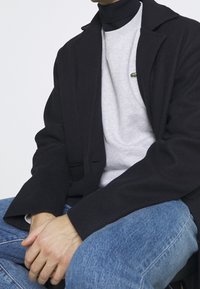Cappotto di lana nero sopra una felpa grigia con un piccolo logo verde, abbinato a jeans di denim blu. Mani visibili appoggiate sulle ginocchia.