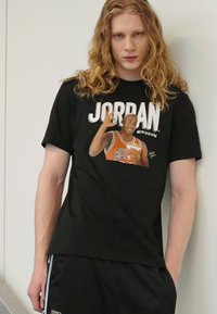 Fekete pamut póló, amelyen egy kosárlabdázó grafika található, a "JORDAN" felirattal a játékos feje fölött, és a "BULLS" narancssárga színben a mezén.