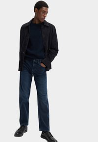 Svart mockajacka över en mörk stickad tröja, tillsammans med mörkblå raka jeans och svarta snörskor.