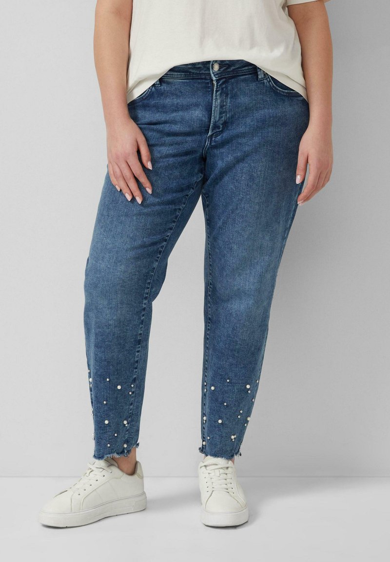 Osoba nosząca niebieskie dżinsy z perełkowymi zdobieniami przy postrzępionych brzegach oraz białe trampki, stojąca na jednolitym tle.