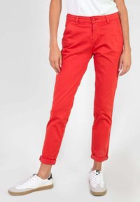 Chino en coton rouge, coupe ajustée avec une fermeture à bouton, manches retroussées et deux poches avant. Porté avec des baskets blanches et un haut léger.