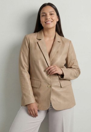 Młoda kobieta uśmiechająca się, ubrana w beżową skórzaną marynarkę i jasne spodnie, stojąca na tle jednolitego jasnoszarego tła.