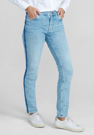 Šviesiai mėlyni liesi džinsai, pagaminti iš džinsinio audinio, su kontrastinga mėlyna šonine juostele, penkiomis kišenėmis ir mygtuko užsegimu, derinami su baltais sportiniais batais.