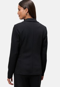 Blazer nero con un design strutturato, colletto a serratura, maniche lunghe e una texture liscia. Presenta una cucitura centrale sul retro.