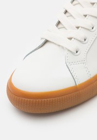 Gros plan d'une basket blanche avec des lacets blancs et une semelle en caoutchouc marron texturée sur un fond clair.