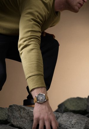 Montre-bracelet en argent avec un cadran foncé, un bracelet en cuir marron et des accents métalliques. La personne qui la porte est en chemise à manches longues olive, accroupie sur des rochers.