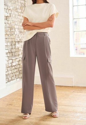 Femme debout, les bras croisés, portant un haut beige à manches courtes, un pantalon cargo taupe à jambes larges et des sandales crème à lanières dans une pièce lumineuse.