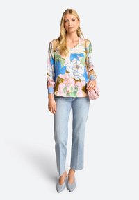 Frau trägt eine mehrfarbige Blumenbluse, hellblaue Jeans, graue spitze Absatzschuhe, goldene Hoops und trägt eine rosa Handtasche mit einer Kettenriemen.
