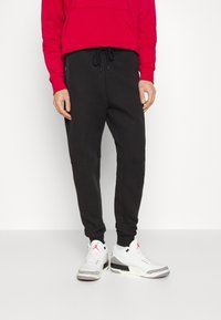 Schwarze Jogginghose mit schmal zulaufendem Bein, seitlichen Reißverschlusstaschen und elastischem Bund mit Kordelzug, kombiniert mit weißen und grauen Sneakers. Rote Kapuzenjacke.