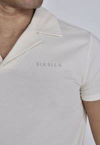Kremfarvet kortærmet polo med en blød tekstur, som har en spids krave og et subtilt "SIKSILK" logo i en lysere nuance.