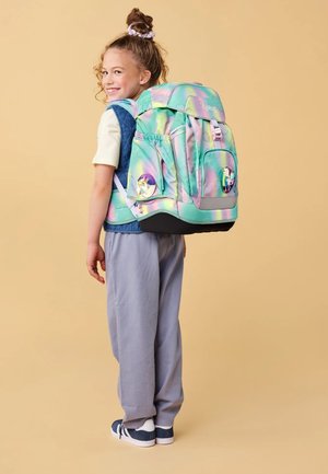 Mochila en colores pastel iridiscentes con acentos gráficos. Múltiples bolsillos y correas acolchadas. Llevada por un niño con atuendo casual, pantalones grises.