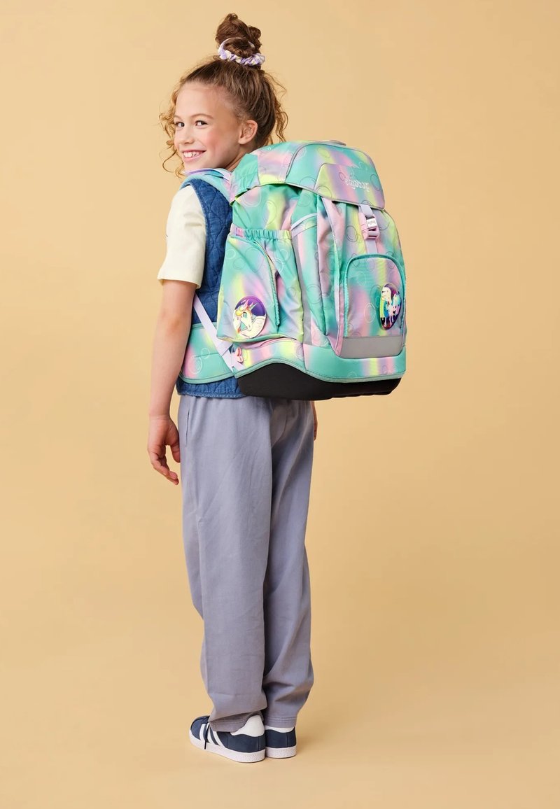 Mochila en colores pastel iridiscentes con acentos gráficos. Múltiples bolsillos y correas acolchadas. Llevada por un niño con atuendo casual, pantalones grises.