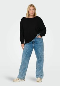 Black oversized knit sweater with a wide neckline, paired with light blue baggy jeans. Model stands with one hand on her hip, neutral background.