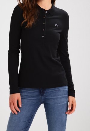 Femme portant un polo noir à manches longues avec des boutons et un petit logo sur la poitrine, associé à un jean bleu.