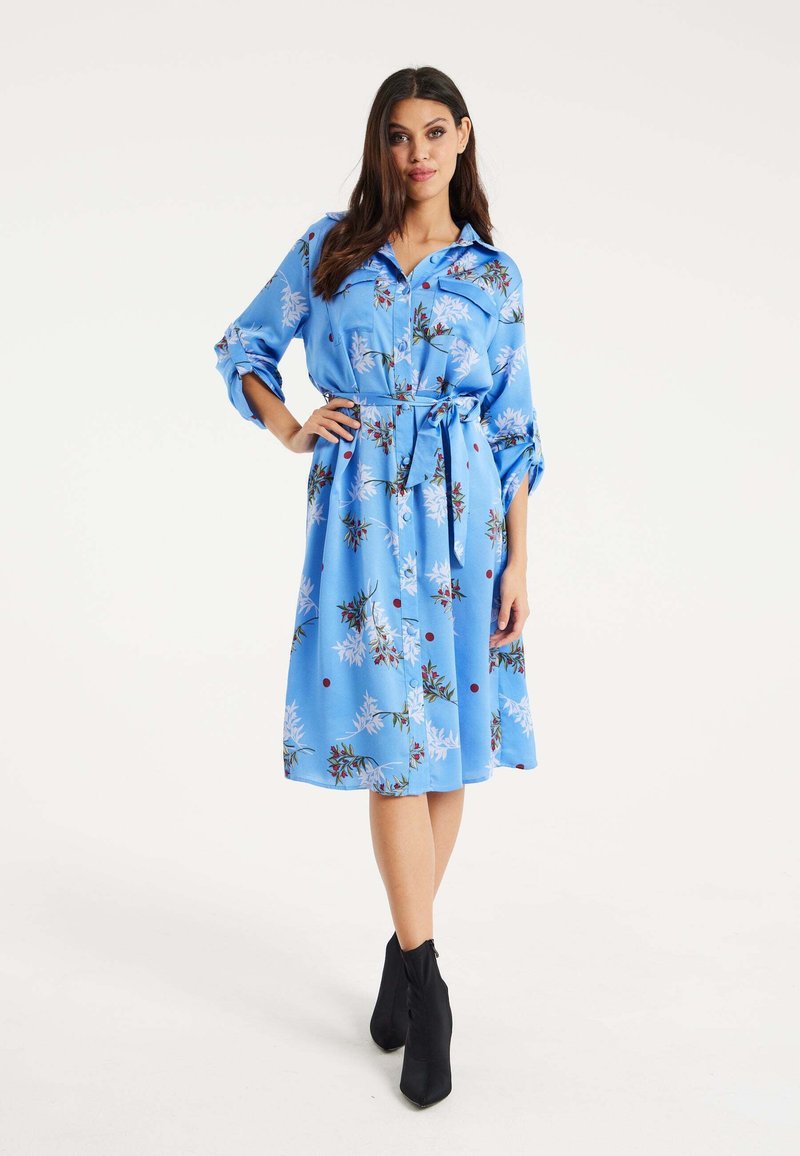 Blue floral shirt dress with a belt, featuring short rolled sleeves, a collar, and a mid-calf length. Black high-heeled boots complete the look.