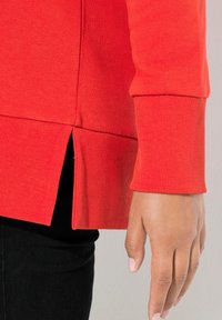 Sudadera roja con puños acanalados y aberturas en los lados del dobladillo, hecha de una tela suave. El diseño incluye un corte ajustado y una textura lisa.