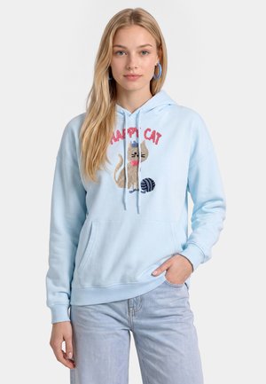 Jonge vrouw draagt een lichtblauwe hoodie met een kattenafbeelding en de tekst "Happy Cat", gecombineerd met lichtblauwe spijkerbroek en blauwe oorringen.