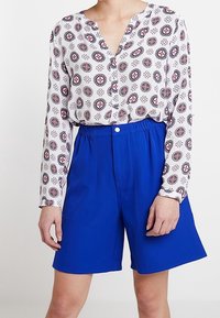 Weißes Langarmbluse mit einem geometrischen roten und blauen Muster, kombiniert mit hellblauen Shorts mit elastischem Bund und Taschen vorn.