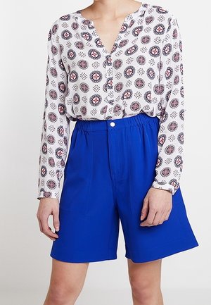 Camicia bianca a maniche lunghe con un motivo geometrico rosso e blu, abbinata a pantaloni corti blu vivaci con vita elastica e tasche frontali.