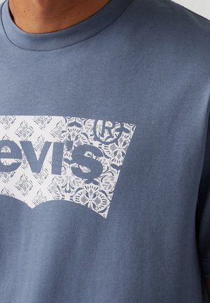 Niebieska koszulka z białym prostokątnym logo na piersi, na którym widnieje słowo "Levi's" w niebieskiej, wzorzystej czcionce.