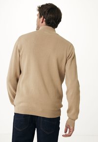 Bežový pletený svetr s vysokým límcem a žebrovanými manžetami. Tkanina má hladkou texturu a je spárována s tmavými džínami.