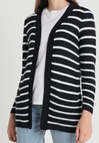 Paski kardigan w granatowo-białe paski, z otwartym przodem, długimi rękawami, o fakturowanej dzianinowej tkaninie, z dwoma przednimi kieszeniami. Noszony na białej bluzce.