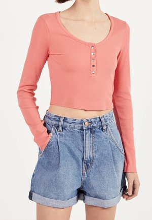 Femme portant un crop top côtelé rose à manches longues avec des boutons en métal et un short en denim taille haute avec des revers et des poches.