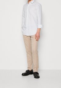 Persoon in een wit gestreept overhemd met lange mouwen, beige broek en zwarte leren schoenen, staande tegen een effen lichte achtergrond.