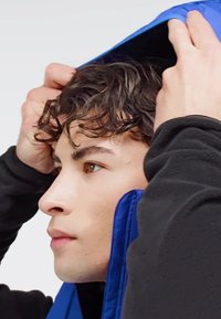 Blå zip-jacka med huva, mjuk textur och kontrasterande svart fleecefoder. Fokusera på ansiktsprofil och händer som justerar huven.