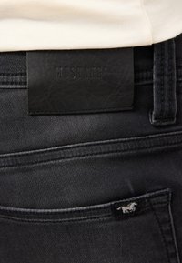 Czarne dżinsy z denimu z matową czarną skórzaną metką z wytłoczonym napisem "MUSTANG" oraz małym logo konia na tylnej kieszeni. Gładka tekstura i dopasowany krój.