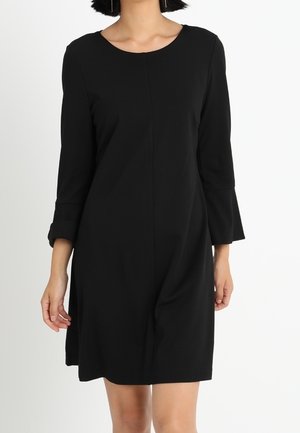 Femme portant une robe noire simple à manches trois-quarts et col rond, debout devant un fond blanc uni.
