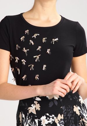 Schwarzes kurzärmeliges Oberteil mit verstreuten rosa und goldenen Perlenverzierungen, kombiniert mit schwarzem und weißem floralen Muster am unteren Teil.