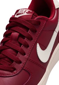 Nike-sneakers hebben een bovenwerk van bordeauxrood leer met witte accenten, een voering van gestructureerde stof en platte bijpassende veters. Opvallend logo op de tong.