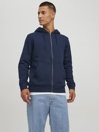 Sudadera de cremallera en color navy, fabricada en una mezcla de algodón, con cordones ajustables, bolsillos frontales y puños acanalados. Combinada con unos vaqueros de mezclilla azul claros.