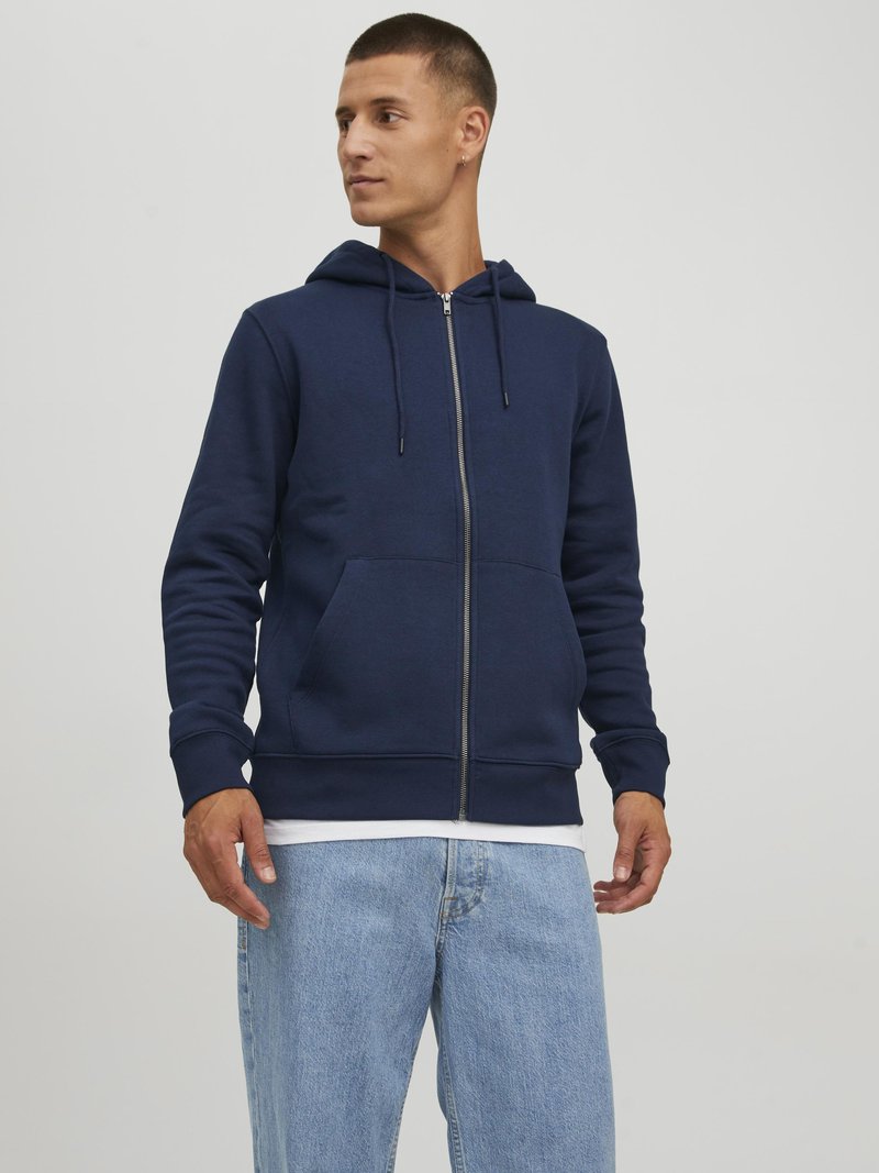 Sudadera de cremallera en color navy, fabricada en una mezcla de algodón, con cordones ajustables, bolsillos frontales y puños acanalados. Combinada con unos vaqueros de mezclilla azul claros.