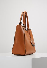 Brązowa skórzana torebka z podwójnymi uchwytami i ozdobnym serduszkiem, umieszczona na białym piedestale na jednolitym tle.