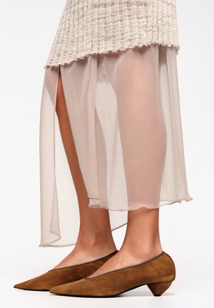 Zapatos marrones de ante con punta afilada y tacón cónico bajo, combinados con una falda traslúcida beige claro con aberturas laterales y dobladillo festoneado.