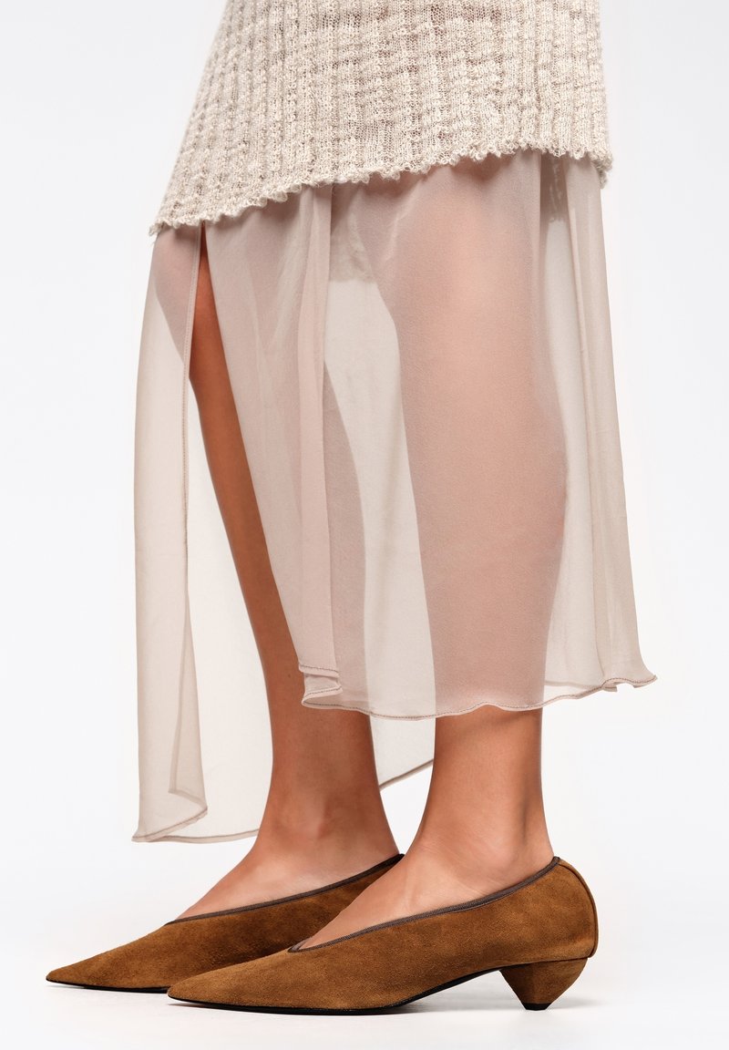 Zapatos marrones de ante con punta afilada y tacón cónico bajo, combinados con una falda traslúcida beige claro con aberturas laterales y dobladillo festoneado.