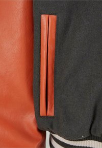 Veste en laine noire avec une poche verticale marron bordée de cuir et un ourlet côtelé rayé noir et blanc.