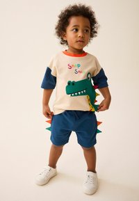Toddler in a cream shirt with a green dinosaur graphic and blue shorts with colorful dinosaur spikes, wearing white sneakers and standing.